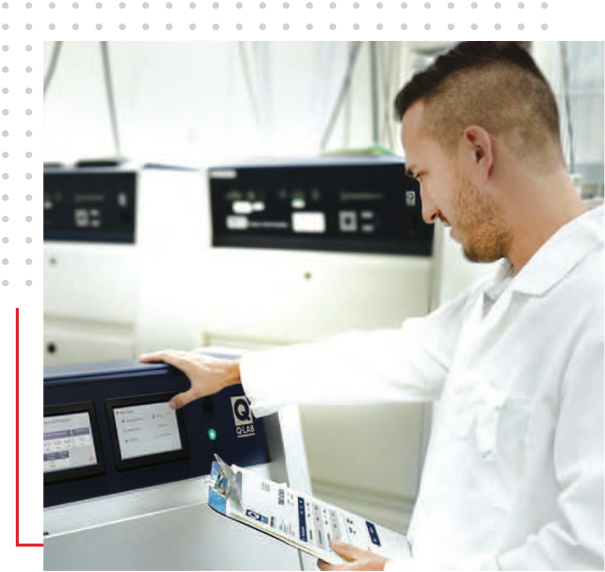 Lab technician in a white coat interacting with the touchscreen controls of Q-Lab equipment, while holding a clipboard with documents.